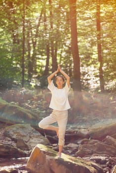 Young woman with closed eyes practicing yoga in pose of tree on stone in forest outdoor.
