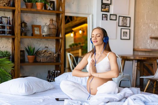 Health care concept. Photo of a young pregnant woman sits on a bed wearing headphones and listens to music for meditation through an online smartphone application.