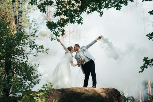 The bride and groom stand on a cliff and hold smoke bombs in their hands in white.