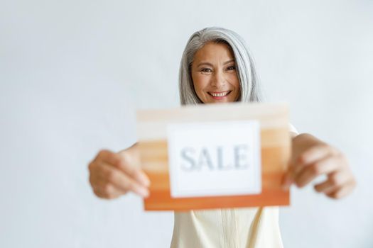 Happy mature Asian woman with long hoary hair shows Sale sign standing on light grey background in studio