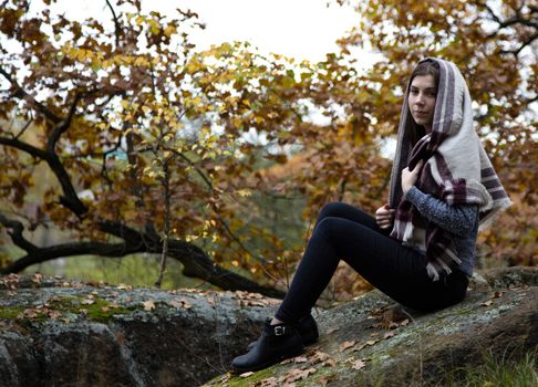 A cute, beautiful, modest religion girl with a scarf or scarf on her head is sitting in the autumn in a park on a stone. Moss and yellow leaves around her.