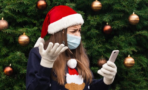 Christmas online greetings. Close up Portrait of woman wearing a santa claus hat and medical mask with emotion. Against the background of a Christmas tree. Coronavirus pandemic