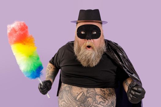 Excited mature man with overweight in Zorro suit with colorful dust brush poses standing on purple background in studio