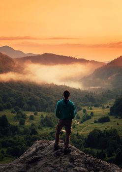 Unrecognizable young man standing on peak of cliff in summer mountains at sunset and enjoying view of nature, rear view.