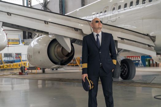 Pilot in a black suit holding a cap in his hands and looking to the side in a hangar