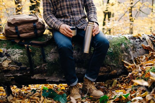 Unrecognizable backpacker young man sitting on fallen tree trunk with cup and thermos and resting in autumn forest.