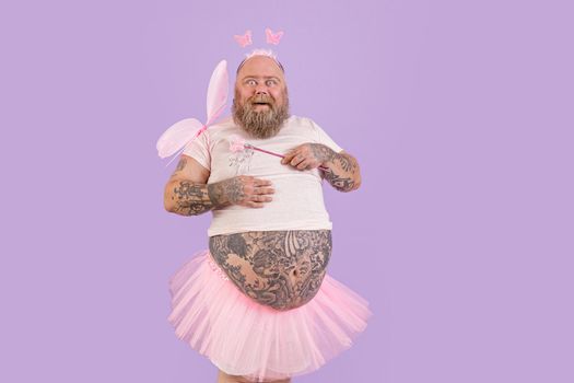 Funny cheerful bearded man with overweight wearing fairy suit with magic stick and wings poses on purple background in studio