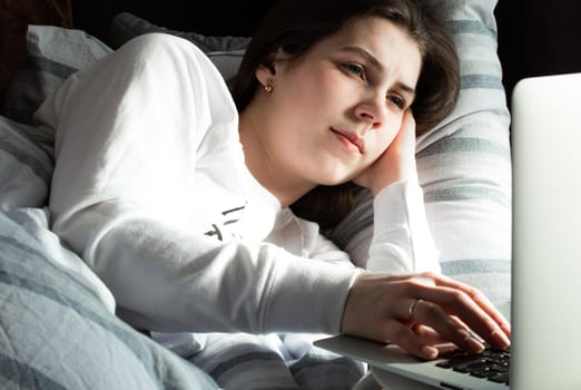 Young cute girl on a sunny day in a white jacket lies on the bed and looks into the laptop.