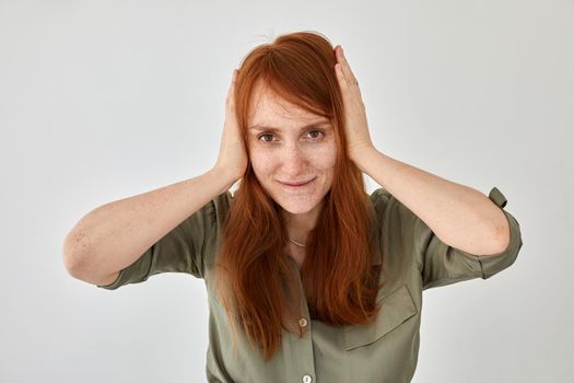 Smiling female with long ginger hair covering ears with hands from noise against white background and looking at camera