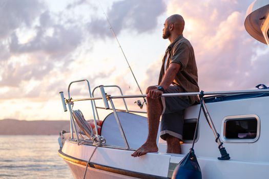 Young african american man standing with fishing rod on a sailboat fishing in open sea on sunset, close up