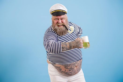 Expressive happy plus size man in sailor suit with smoking pipe holds glass mug of delicious beer on light blue background in studio
