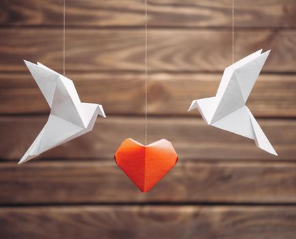 Two origami dove birds around red paper heart on wooden background. Greeting card for Valentines day.