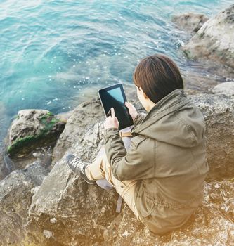 Young woman sitting on stone coast near the sea and working on digital tablet.