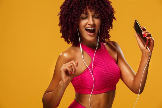 Beautiful young and fit woman in headphones listening music from phone and smiling with closed eyes standing on a yellow background in a pink sportswear