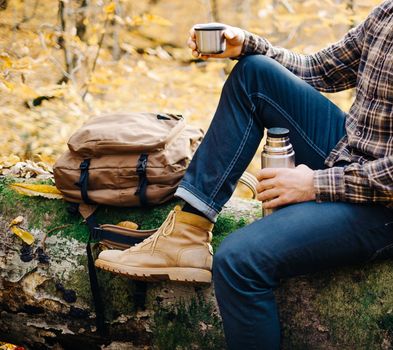 Unrecognizable backpacker young man resting on fallen tree trunk with cup and thermos in autumn forest.