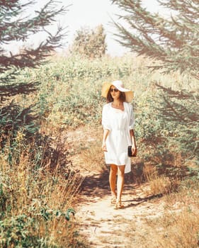 Stylish beautiful young woman in yellow dress and hat with wide brim walking on footpath in summer park.