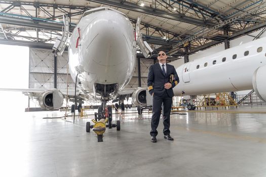 Serious pilot posing in black suit and sunglasses near plane in airport hangar