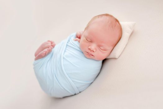 Portrait of newborn baby boy swaddled in light blue fabric sleeping on white background and holding tiny feet up. Adorable infant child napping during studio photoshoot
