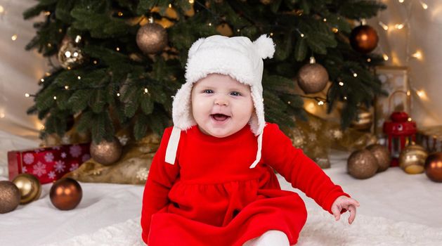 Christmas baby is smiling. A cute little girl in a red dress and white hat expresses emotions. Christmas concept with little kid, tree and garland on background in blur