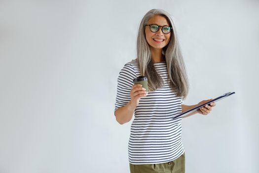 Cheerful mature Asian lady with natural silver hair holds clipboard and cup standing on light grey background in studio, space for text