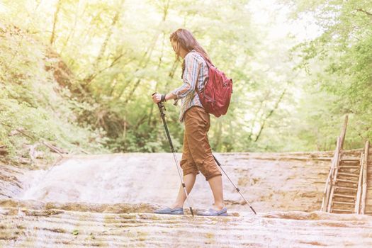 Backpacker young woman crossing a river with trekking poles on sunny summer day.