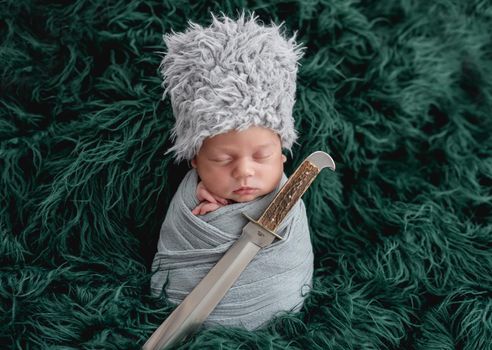 Newborn in ethnic hat sleeping with dagger on green background