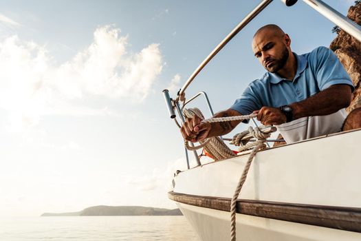 Young african american sailor tying ropes on sailboat in the sea on sunset, close up