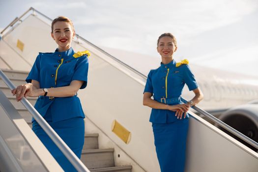 Two beautiful air stewardesses in blue uniform smiling at camera, standing on airstair and welcoming passengers. Aircrew, occupation concept