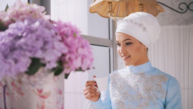 Beautiful muslim model in blue wedding dress drinking coffee near the window, close up