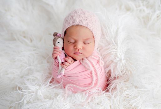 Swaddled beautiful newborn baby girl sleeping in white fur and holding knitted doll in her hands. Cute female infant child photoshoot in studio