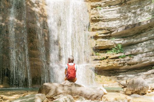 Traveler young woman with backpack sitting on stone in front of waterfall in summer outdoor.