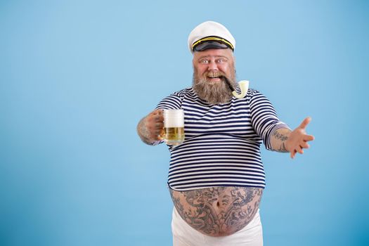 Joyful bearded obese person in sailor costume with smoking pipe holds glass of fresh foamy beer standing on light blue background in studio