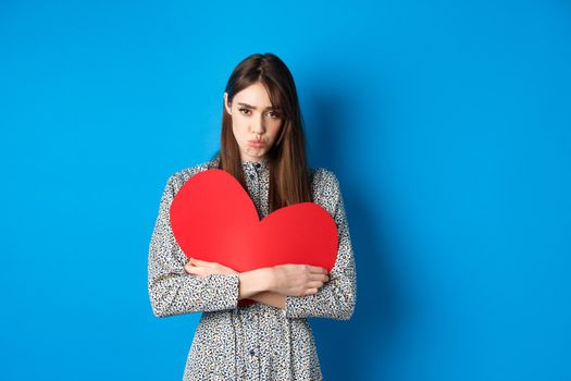 Valentines day. Sad and gloomy caucasian girl pucker lips and looking disappointed, holding big red heart cutout, standing on blue background.