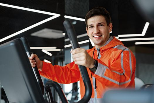 Portrait of a young man in orange windbreaker workout on a fitness machine at a gym.