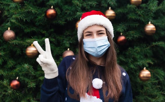 Christmas piece. Close up Portrait of woman wearing a santa claus hat and medical mask with emotion. Against the background of a Christmas tree. Coronavirus pandemic