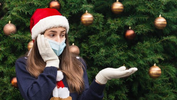 Christmas without a gift. Close up Portrait of woman wearing a santa claus hat and medical mask with emotion. Against the background of a Christmas tree. Coronavirus pandemic
