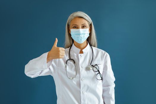 Positive mature Asian female doctor in uniform with surgical mask and stethoscope shows thumb up on blue background in studio. Professional medical staff