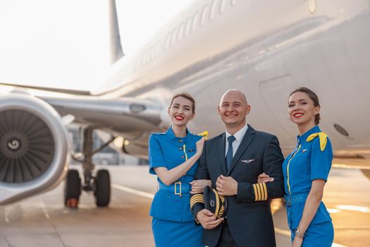 Portrait of excited male pilot posing together with two air hostesses in blue uniform in front of an airplane in airport. Aircraft, aircrew concept