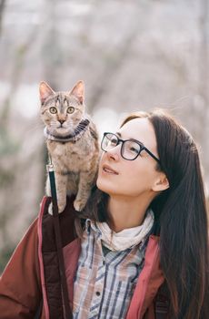 Traveler cat wearing in bandana sitting on shoulder of young woman outdoor.