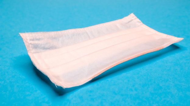 One 3-ply disposable surgical face mask with rubber grips covering mouth and nose against blue background. Protection from bacteria, healthcare and medicine concept. COVID-19. A dangerous virus
