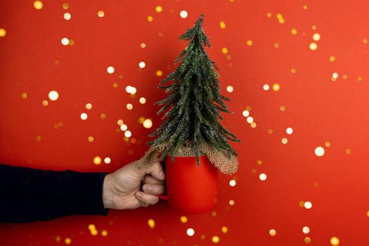 Caucasian man hand holding a red mug with pine tree, symbol of Christmas and new year, creative Christmas concept, red bokeh background, copy space.