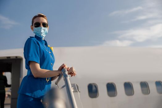Portrait of air stewardess in blue uniform, sunglasses and protective face masks looking at camera, standing on airstair on a daytime. Aircrew, occupation, covid19 concept