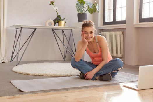 Smiling athletic female in sportswear sitting on mat with laptop during online workout at home and looking at camera