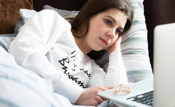 Young cute girl on a sunny day in a white jacket lies on the bed and looks into the laptop.