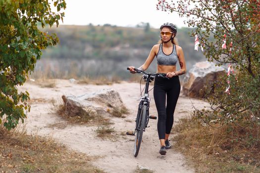 Fit young woman wearing sportswear standing with her bicycle on rocky background at autumn day