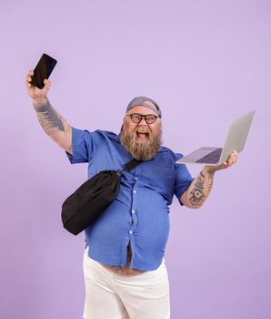 Positive bearded plus size man with laptop and crossbody bag shows mobile phone with blank screen standing on purple background in studio