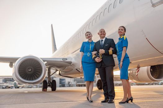 Full length shot of excited male pilot posing together with two air hostesses in blue uniform in front of an airplane in terminal at sunset. Aircraft, aircrew concept