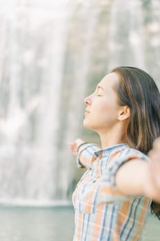 Young woman with closed eyes meditating with raised arms on background of waterfall outdoor, side view.