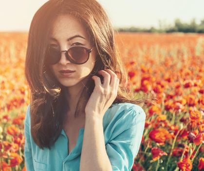 Attractive brunette young woman in sunglasses walking in spring poppies flowers meadow, looking at camera.