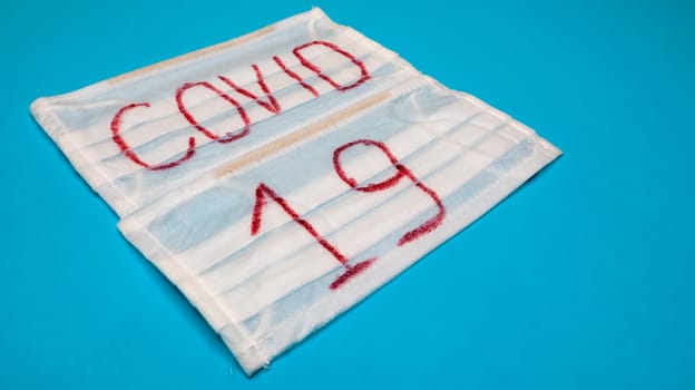 Typical white 3-ply surgical mask with rubber earhooks to cover mouth and nose with English block letters Covid-19 on a blue background. Dangerous virus and quarantine concept